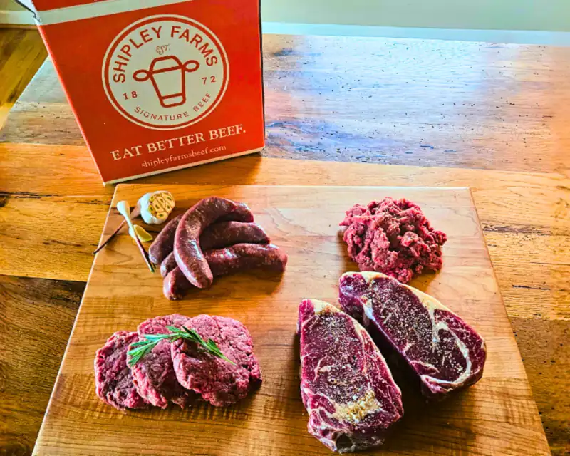A Shipley Farms box beside a cutting board of ribeye steaks, sausage links, ground beef and burger patties