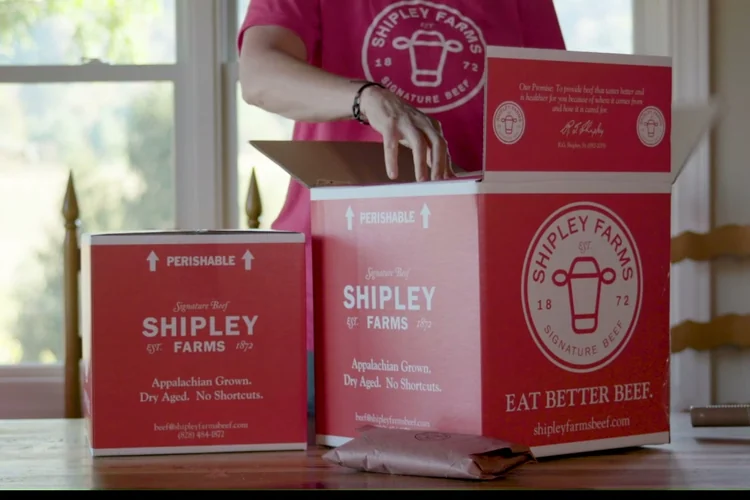 A person opening a red Shipley Farms shipping box of dry-aged beef on a kitchen counter, with a second sealed box beside it